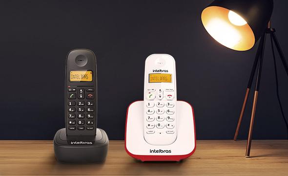 Imagem de dois telefones com suporte de telefones sem fio, preto e branco, com uma lâmpada iluminando os fois em cima de uma mesa